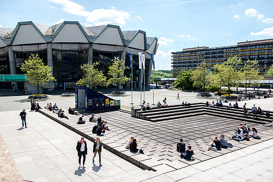 Photo: A photo of the sunny RUB campus with students and the main lecture hall.