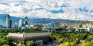 Photo: An overview of the Kazakh capital Almaty showing modern glass buildings, a blue cloudy sky, lush trees and and snow-capped mountains in the background.