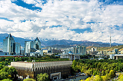 Foto: Ein Blick auf die kasachische Hauptstadt Almaty mit modernen Glasgebäuden, einem blauen, bewölkten Himmel, sattgrünen Bäumen und schneebedeckten Bergen im Hintergrund.