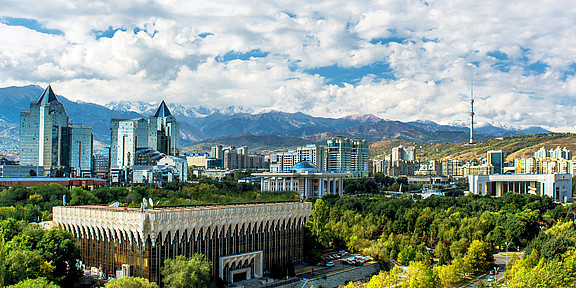 Photo: An overview of the Kazakh capital Almaty showing modern glass buildings, a blue cloudy sky, lush trees and and snow-capped mountains in the background.