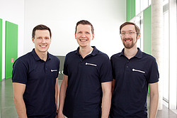 Three young men wearing black polo shirts in the hallway of an office building.