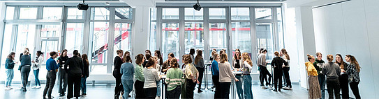 Photo: Female scientists stand in groups around tables in a bright room and chat.