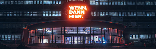 A photo of a building, its entrance, and windows at night. Above the entrance, the words “If, then here” are illuminated.