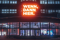 A photo of a building, its entrance, and windows at night. Above the entrance, the words “If, then here” are illuminated.