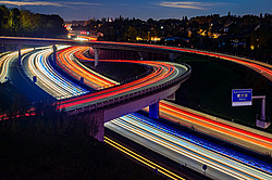Nighttime photograph of a highway interchange in the Ruhr region; the taillights form red stripes, while the headlights form white stripes.