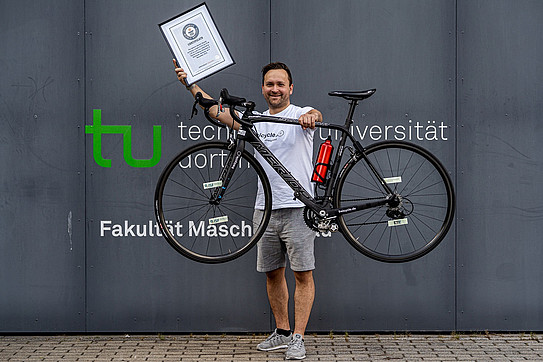 Young man lifts a racing bicycle and holds a certificate in his other hand.