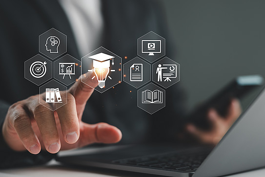 A man sitting at a laptop with a cell phone in his hand presses his right index finger on a graphic overlay showing a light bulb with a doctor's hat.