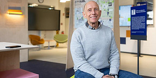 A middle-aged man in a light blue sweater is sitting on a stool in front of a wall covered with posters.