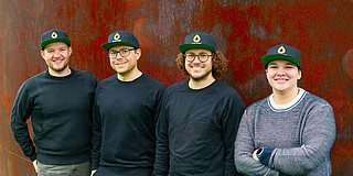 Four people, each wearing the same cap, are standing in front of a rust-brown wall.
