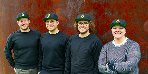 Four people, each wearing the same cap, are standing in front of a rust-brown wall.