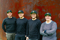 Four people, each wearing the same cap, are standing in front of a rust-brown wall.