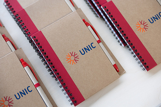 Drei Reihen übereinander sortierte Ringbuchkladden, hellbraun mit rotem Rand, darauf Logo "UNIC"