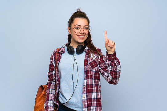 Young woman in a lumberjack shirt, wearing glasses and headphones around her neck, raises the index finger of her left hand.