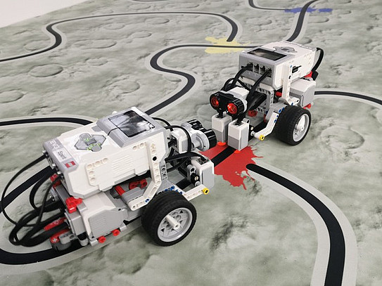 Two homemade robots on wheels drive across a replica of the lunar landscape.