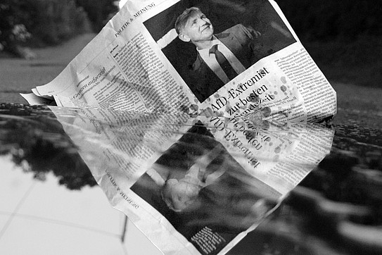 Black-and-white photo: A newspaper with a political article about Björn Höcke lies in a puddle.