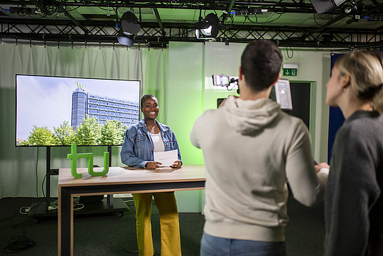 Photo: A student speaks into the camera in a studio. Behind her, a screen shows the Math Tower at TU Dortmund University. Two other students operate the camera.