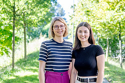 Photo: Two young women smile at the camera. Behind them are trees with green leaves.