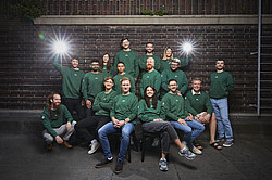 Ein Gruppenfoto des Teams von vGreens in grünen Pullovern vor einer Ziegelsteine.