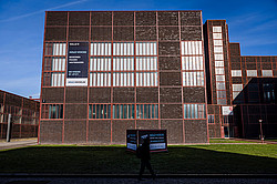 Photo: A large brick building with windows and banners advertising the “HOLO-VOICES” exhibition.