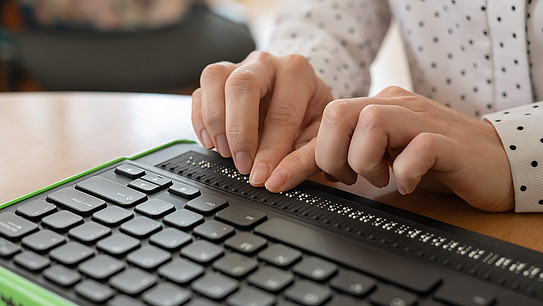 A blind woman uses a computer with a Braille display and a computer keyboard. 