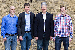 Four men in suits and shirts stand in front of a pile of earth with tire tracks.
