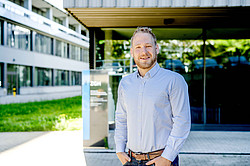 Photo: A man in a light blue shirt stands in front of the ZGH building on the campus of Ruhr University Bochum.