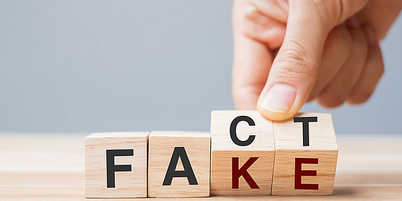 One hand gently turns two dice marked with the letters “C” and “T” so that another side, marked with the letters “K” and “E,” is visible, turning the word ‘Fact’ into “Fake.”