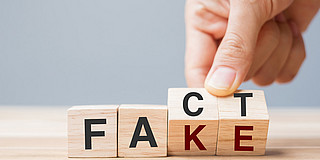 One hand gently turns two dice marked with the letters “C” and “T” so that another side, marked with the letters “K” and “E,” is visible, turning the word ‘Fact’ into “Fake.”