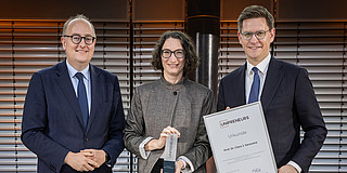 Foto: Auf dem Foto sind drei Menschen zu sehen. Eine Frau bekommt eine Urkunde verliehen. 
