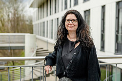 Photo of Prof. Silvana Botti: Prof. Botti is wearing black clothing and leaning against a railing. In the background is a white, modern building.