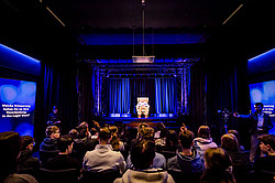 A group of people is sitting in a dark room, watching a hologram of an elderly person on an illuminated stage with an armchair in the center.