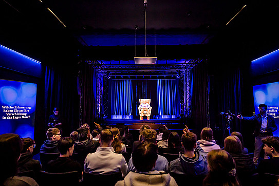 Eine Gruppe von Menschen sitzt in einem dunklen Raum und betrachtet ein Hologramm einer älteren Person auf einer beleuchteten Bühne mit einem Sessel im Zentrum.