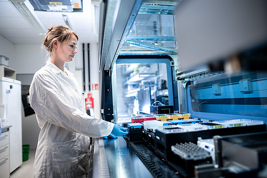 A scientist pushes samples into a modern sampling robot in the laboratory, wearing a lab coat and gloves.