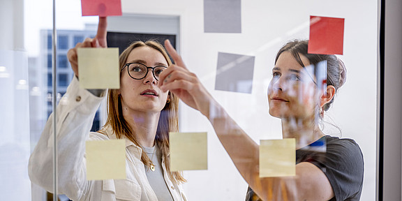 Two women are standing in front of a glass pane covered with Post-its