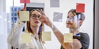 Two women are standing in front of a glass pane covered with Post-its