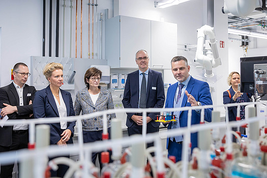 Six people are standing in a laboratory in front of a piece of equipment that is faintly visible in the foreground. A man is explaining the equipment to the group. Among them is Ina Brandes, Minister of Culture and Science for North Rhine-Westphalia.