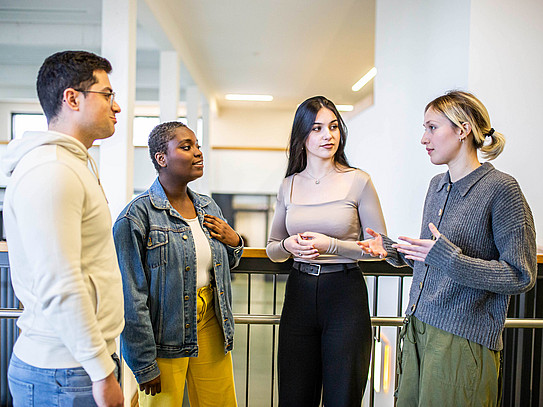 Das Bild zeigt vier Studierende, die in einem Halbkreis zueinander stehen. Drei Personen der Studierenden richten alle den Blick auf eine sprechende Person.