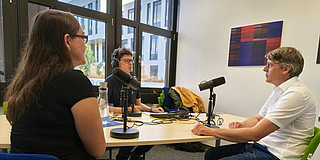 Drei Personen sitzen an einem Tisch und nehmen einen Podcast auf.