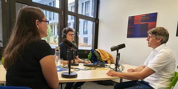Drei Personen sitzen an einem Tisch und nehmen einen Podcast auf.