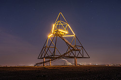 Tetrahedron sculpture illuminated at night in the upper third, industrial backdrop further away in the background.