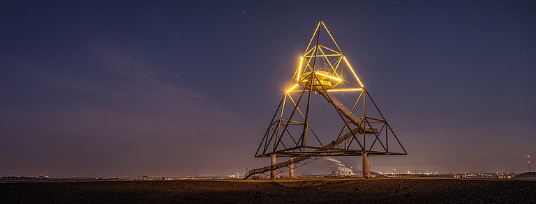 Tetrahedron sculpture illuminated at night in the upper third, industrial backdrop further away in the background.