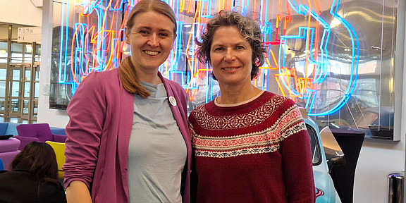 Foto: Prof. Doris Reiter und Dr. Christiane Wüllner stehen in einem Raum vor bunter Leuchtschrift.