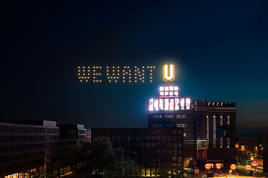 Night shot of the illuminated Dortmunder U, with the words “We want” superimposed next to the "U". 
