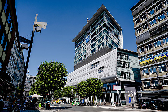 A photo of a street in the city center of Bochum. It shows modern buildings, green trees and a blue sky.