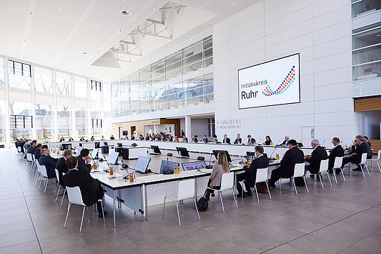 Foyer of a modern exhibition hall, conference participants are seated at long tables arranged in a large rectangle, the logo of the Initiativkreis Ruhr is displayed on the wall.