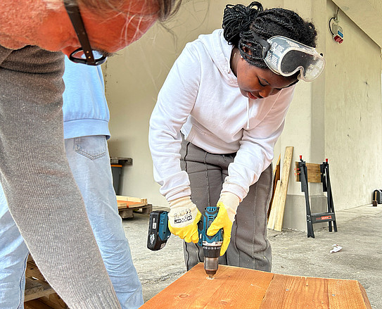 Girl wearing safety goggles and gloves drills a hole in a wooden board with a drill, other people in the foreground