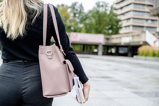 Rear view of a woman with a large, pale pink shoulder bag and books in her hand walking across the RUB campus