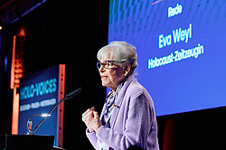 Photo: A person speaks at a podium in front of a large screen with a blue background and text.