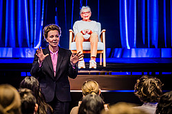 Photo: A person speaks in front of an audience while a hologram is illuminated in blue light on stage.