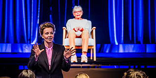 Photo: A person speaks in front of an audience while a hologram is illuminated in blue light on stage.